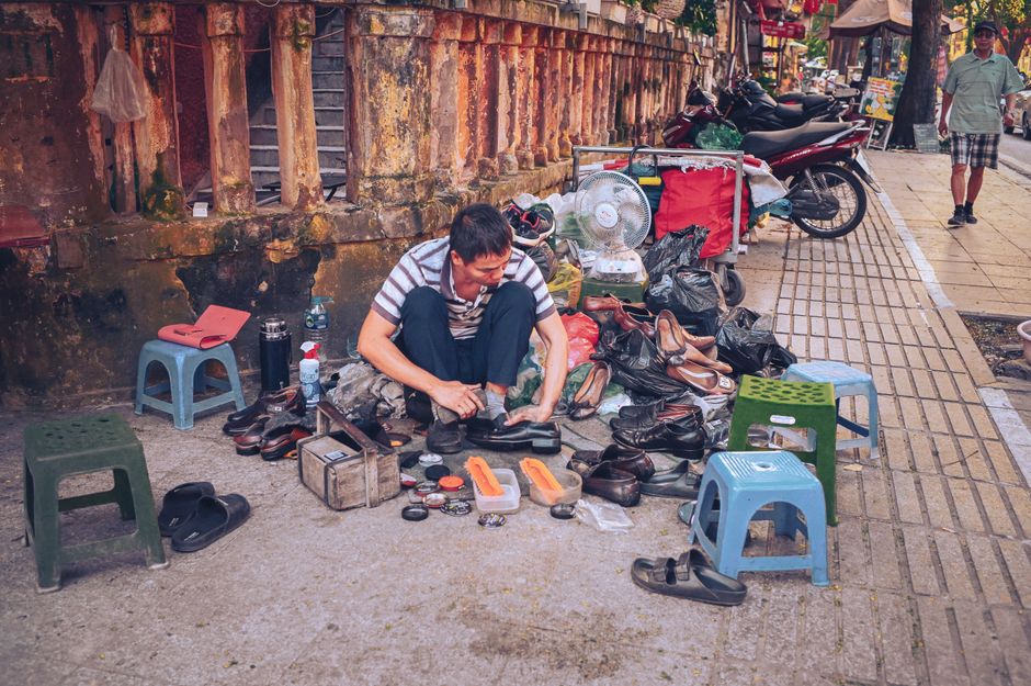 A shoe repairman working on the street surrounded by footwear and tools.