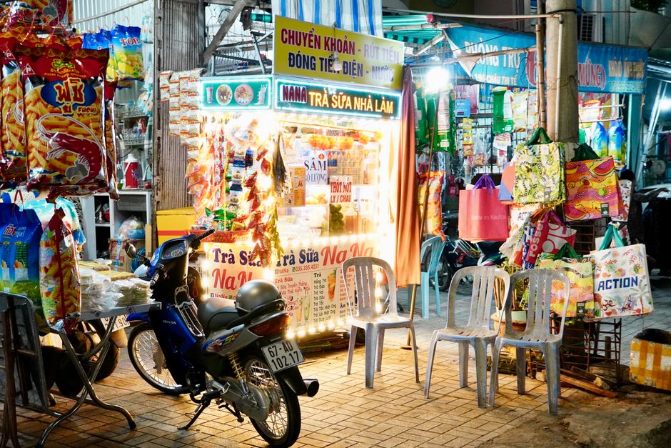 Colorful street vendor stall at night market with hanging snacks and plastic chairs, Vietnam.