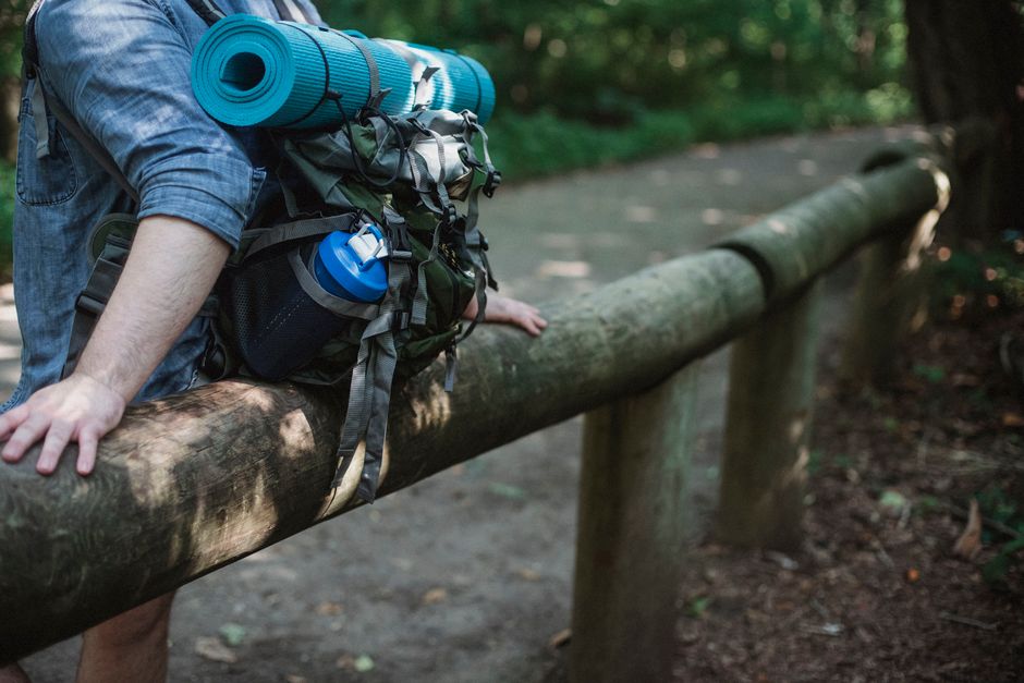Crop unrecognizable male backpacker with equipment for hiking leaning on wooden railing while having break during trekking
