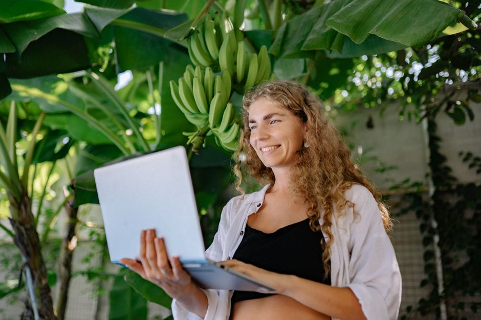 A woman enjoys remote work amidst lush banana plants, smiling at her laptop.