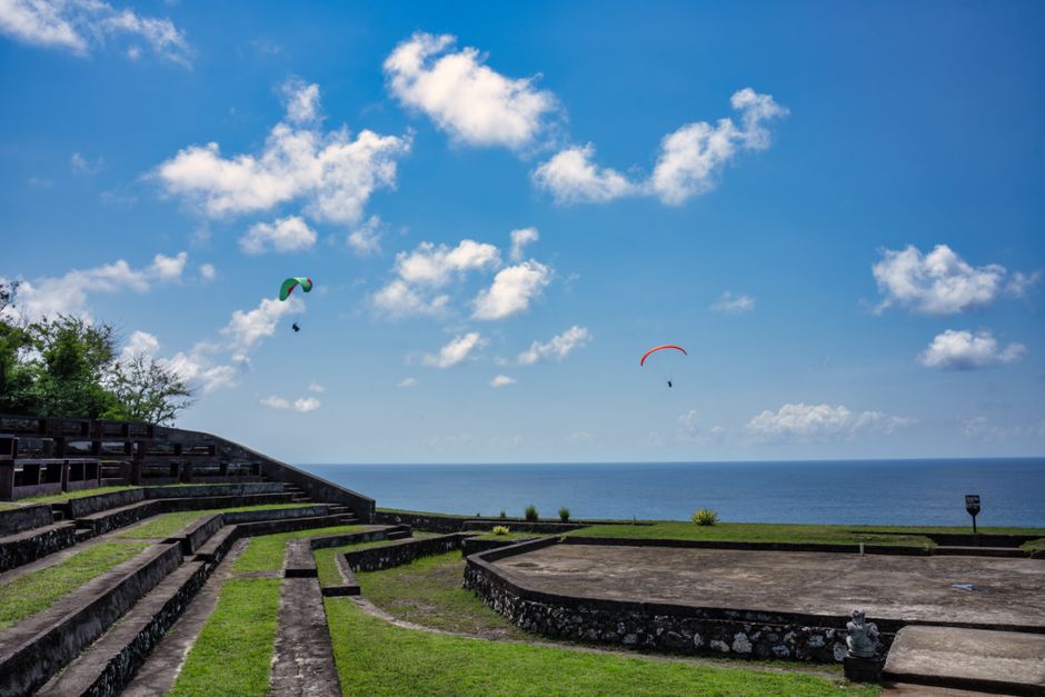 Two paragliders flying over lush greenery and ocean views in Bali, Indonesia, under a bright blue sky.