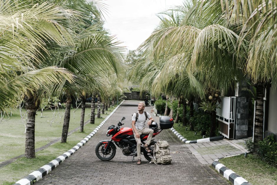 Full length adult male traveler leaning on motorbike and helmet near duffel back while resting on street among palms