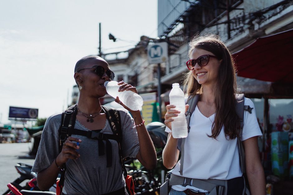 Two women, smiling and holding water bottles, enjoy a sunny day while traveling.