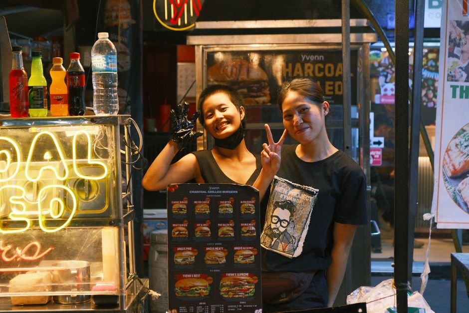 Two smiling street food vendors at a vibrant night market stall selling burgers.