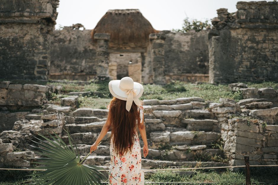 A woman in a floral dress and sun hat explores ancient stone ruins on a sunny day.