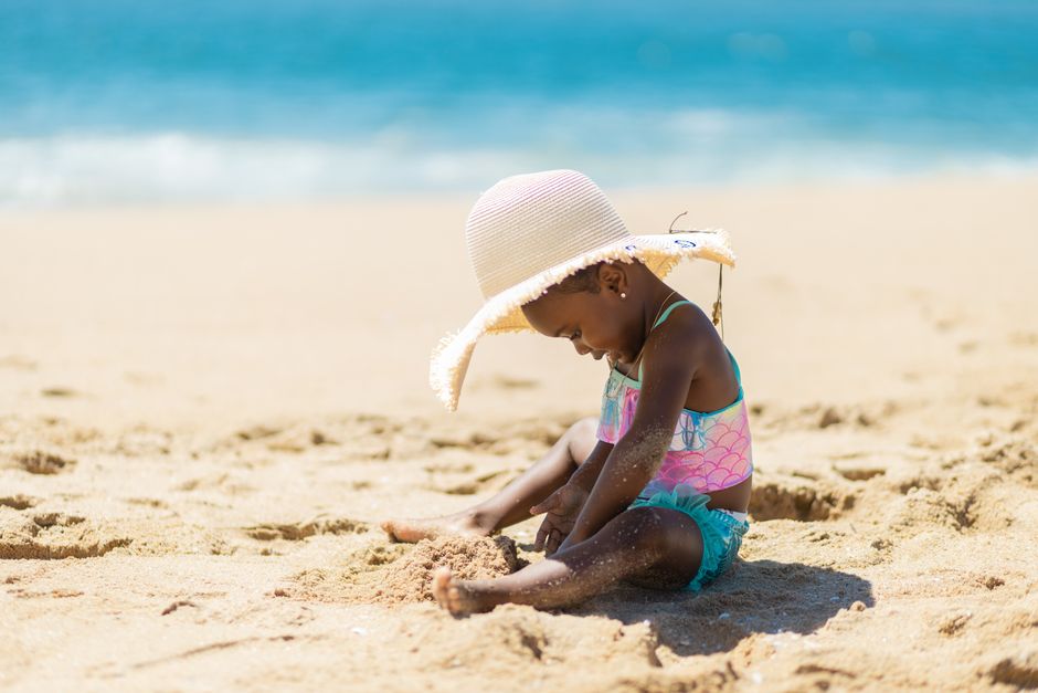 Little girl in sun hat playing on the sandy beach under the summer sun, having fun in swimwear.