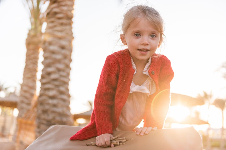Cheerful little girl in trendy red cardigan playing on sunny beach near exotic palms and looking at camera happily at sunset