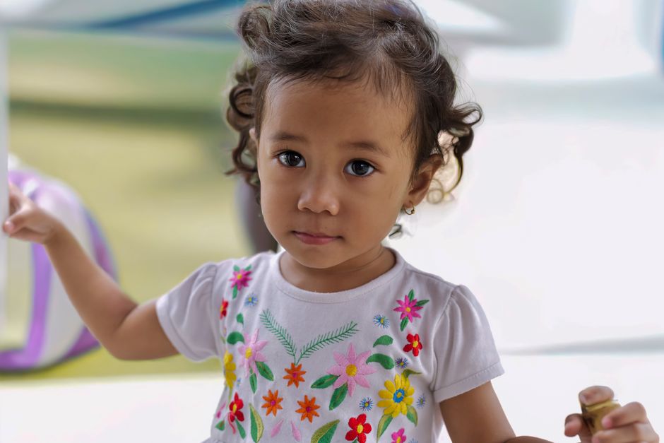 Adorable young girl with curly hair and floral dress looking curiously at the camera indoors.