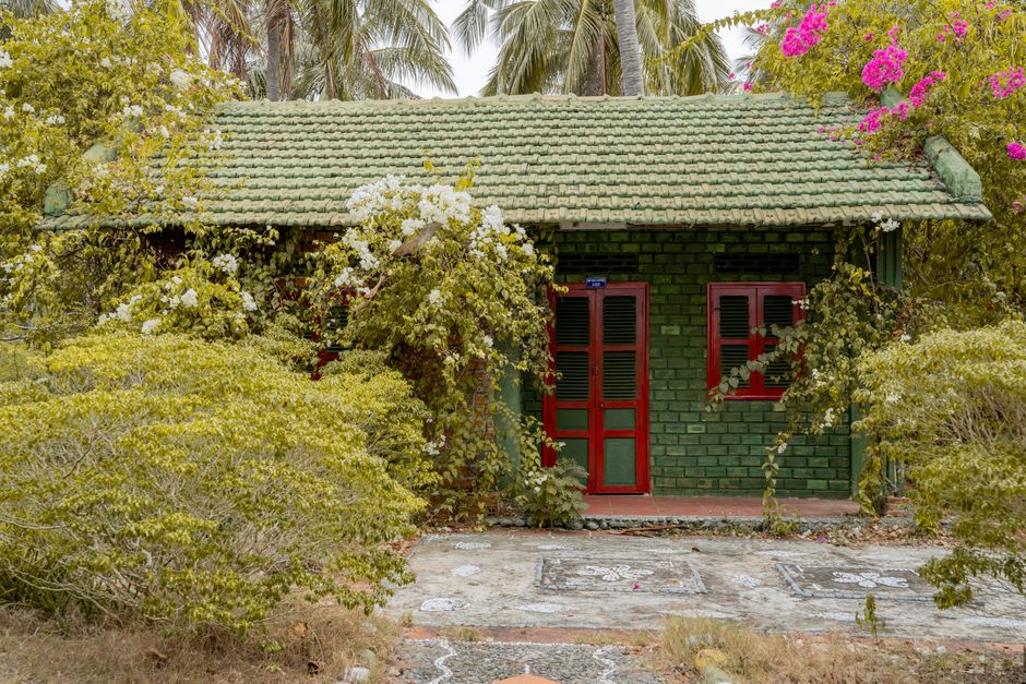 A quaint green cottage with a tiled roof nestled among vibrant flowers and greenery.