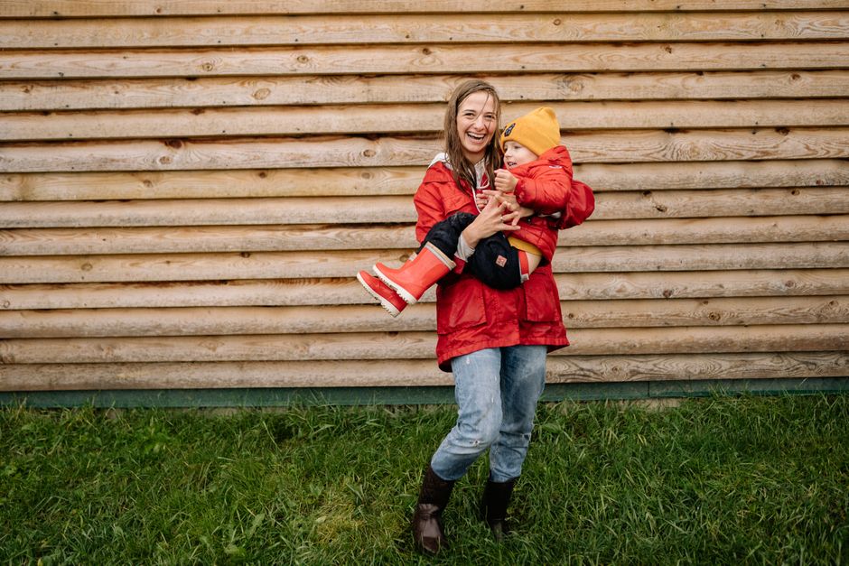 A mother joyfully holds her child against a rustic wooden wall, both in red outfits.