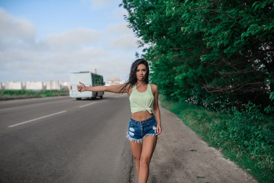 Woman in summer attire hitchhiking on rural road with urban background.