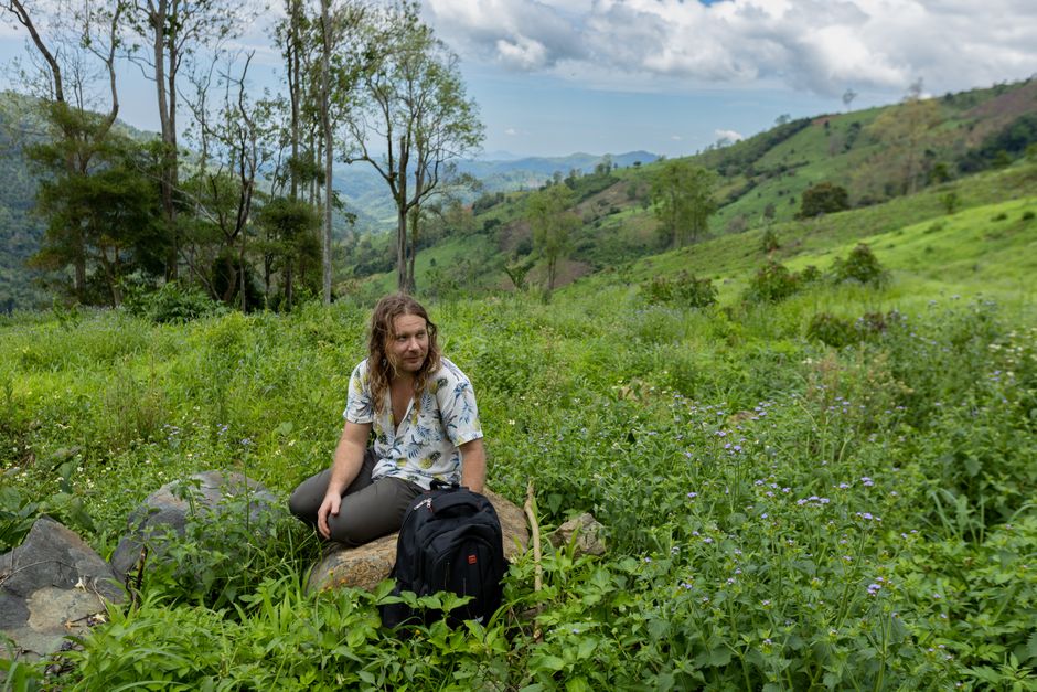 A man with long hair sits on a rocky hillside surrounded by lush greenery and trees.