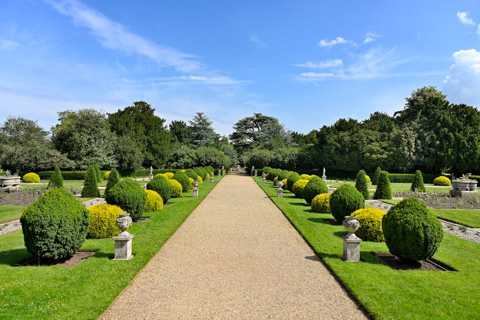 Elegant garden pathway lined with manicured bushes and classic pedestals under a clear sky.