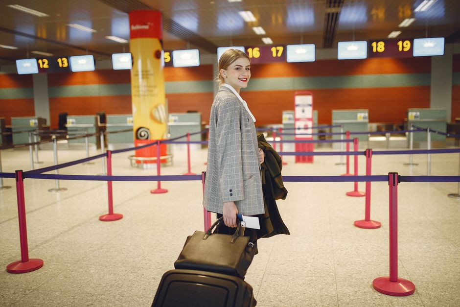 Side view of happy businesswoman in trendy jacket carrying baggage and smiling at camera while waiting for flight in modern airport