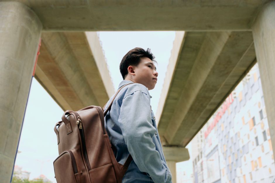 Young man with a backpack gazing upward under an urban bridge, depicting modern city exploration.