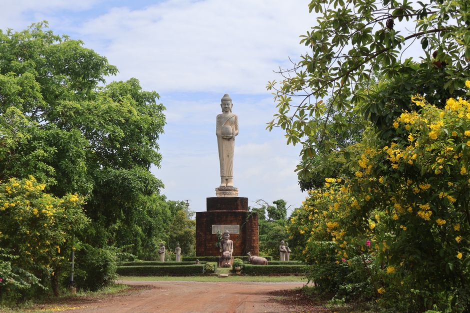 A tall Buddha statue surrounded by lush greenery and vibrant flowers on a sunny day.