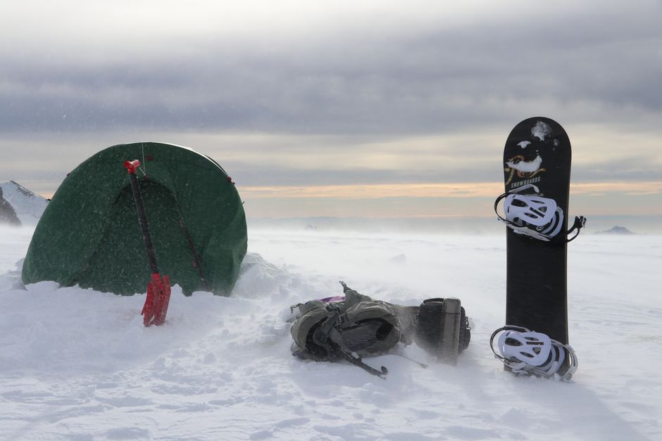 Snowy mountain camping setup with a tent, snowboard, and backpack in winter.