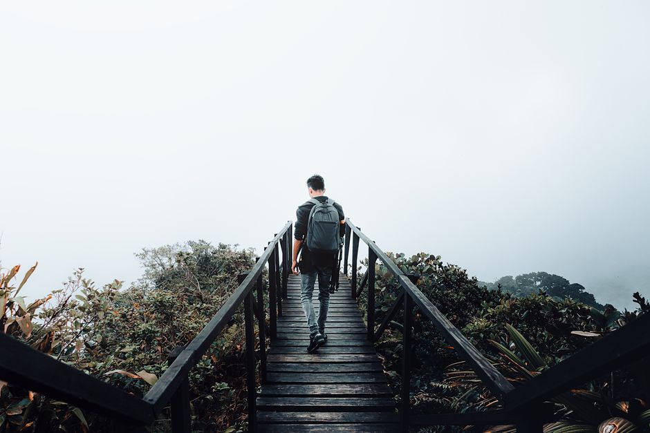 A man with a backpack walks on a wooden path surrounded by misty foliage and mountains.