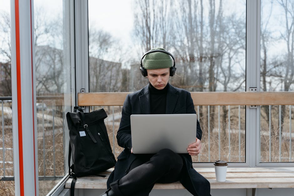 Man sitting at a bus stop working on a laptop, embracing remote work lifestyle.