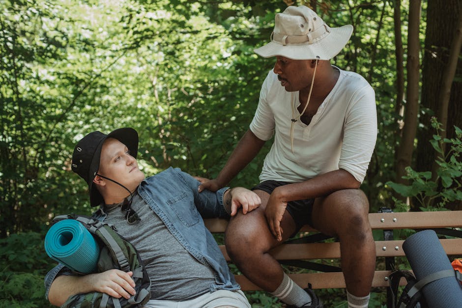 Multiethnic couple of gays in panama hats with hiking equipment looking at each other while resting on bench