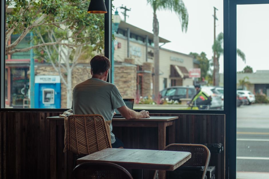 A man working on a laptop in a cafe, overlooking city streets through a large window.