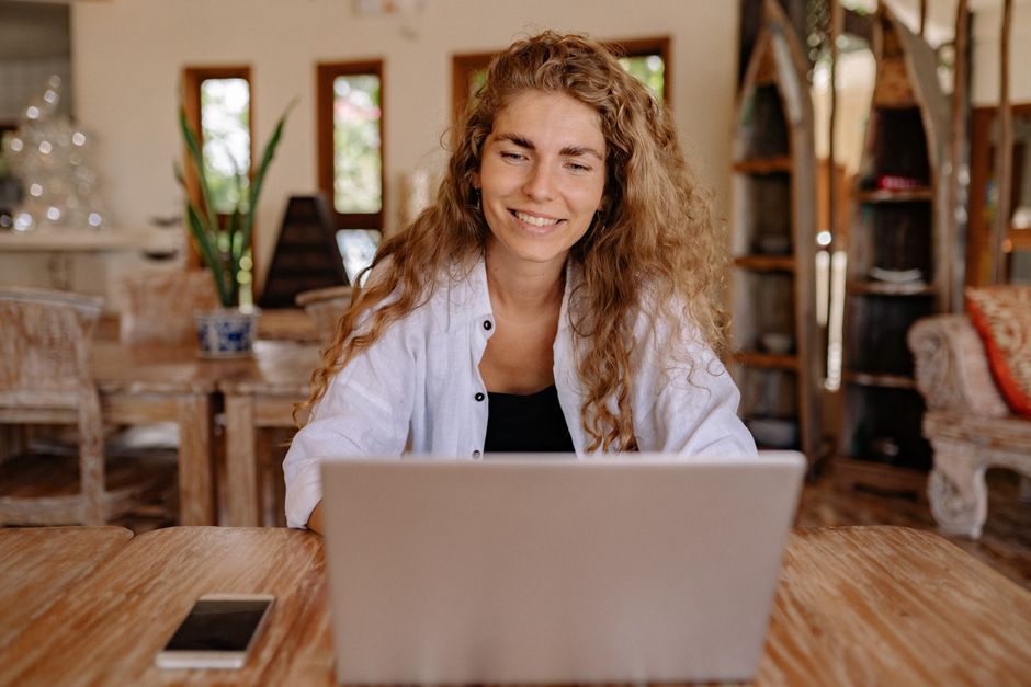 A woman smiling while working on a laptop at a wooden table indoors, embracing remote work lifestyle.